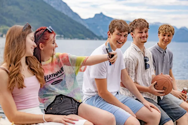 Fünf Schüler:innen sitzen auf der Seemauer, eine macht ein Selfie; im Hintergrund der Traunsee und der Traunstein.