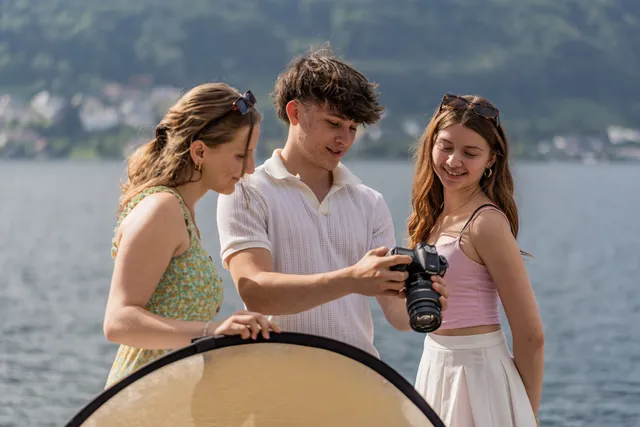 Drei Schüler:innen begutachten am Seeufer gemeinsam das Display einer Spiegelreflexkamera, eine hält einen Reflektor.
