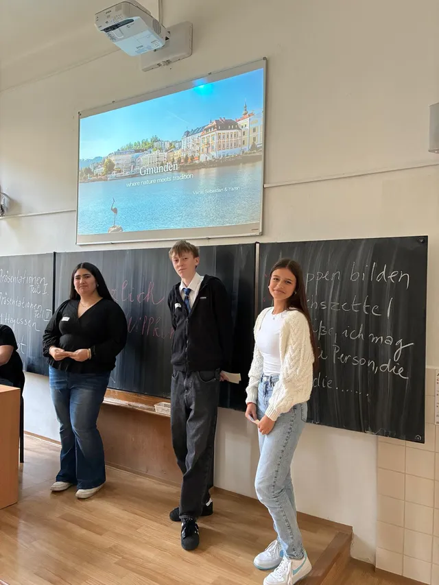 Drei Schüler:innen präsentieren vor der Tafel; auf der Projektionsfläche eine Folie mit dem Titel „Gmunden – where nature meets tradition“.