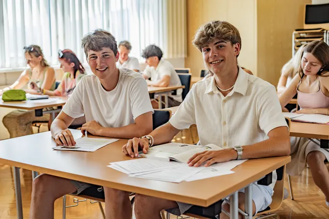 Zwei Schüler sitzen lächelnd nebeneinander am Tisch, vor ihnen Unterlagen und ein aufgeschlagenes Buch.